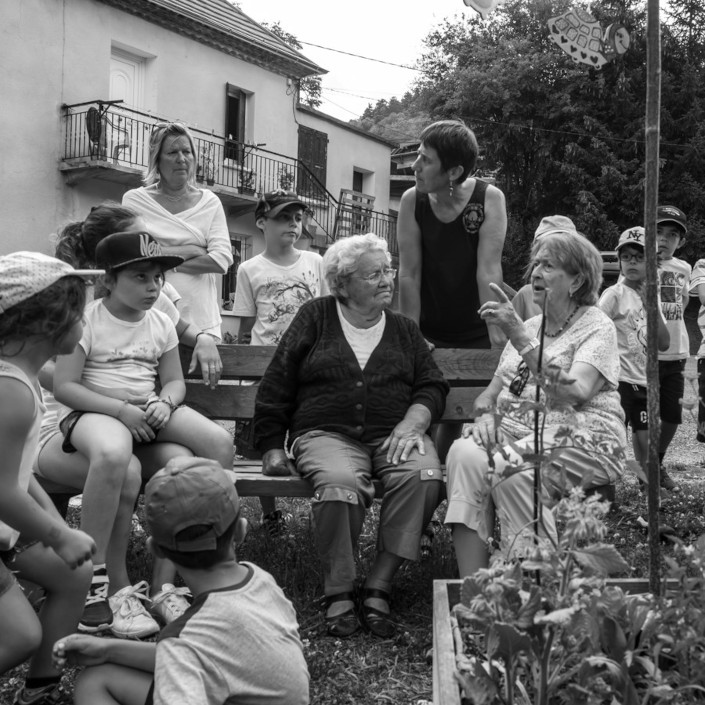 Deux dames âgées discutent sur un banc, autour d'elle des enfants et des femmes les écoutent.