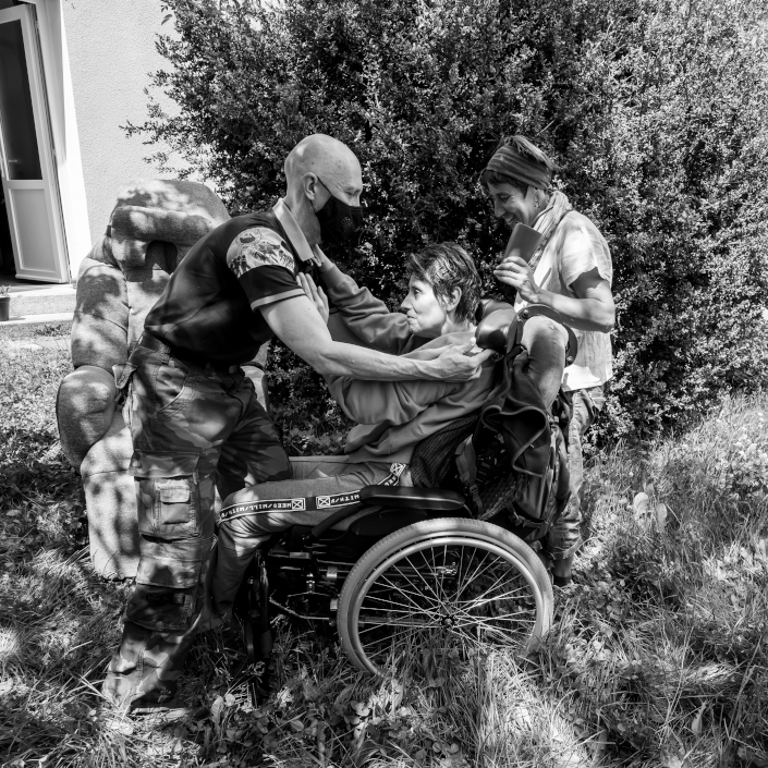 Un bénévole aide une femme à sortir de son fauteuil roulant. Penché vers elle, il la saisit par les épaules avec délicatesse. Derrière, une femme sourit.