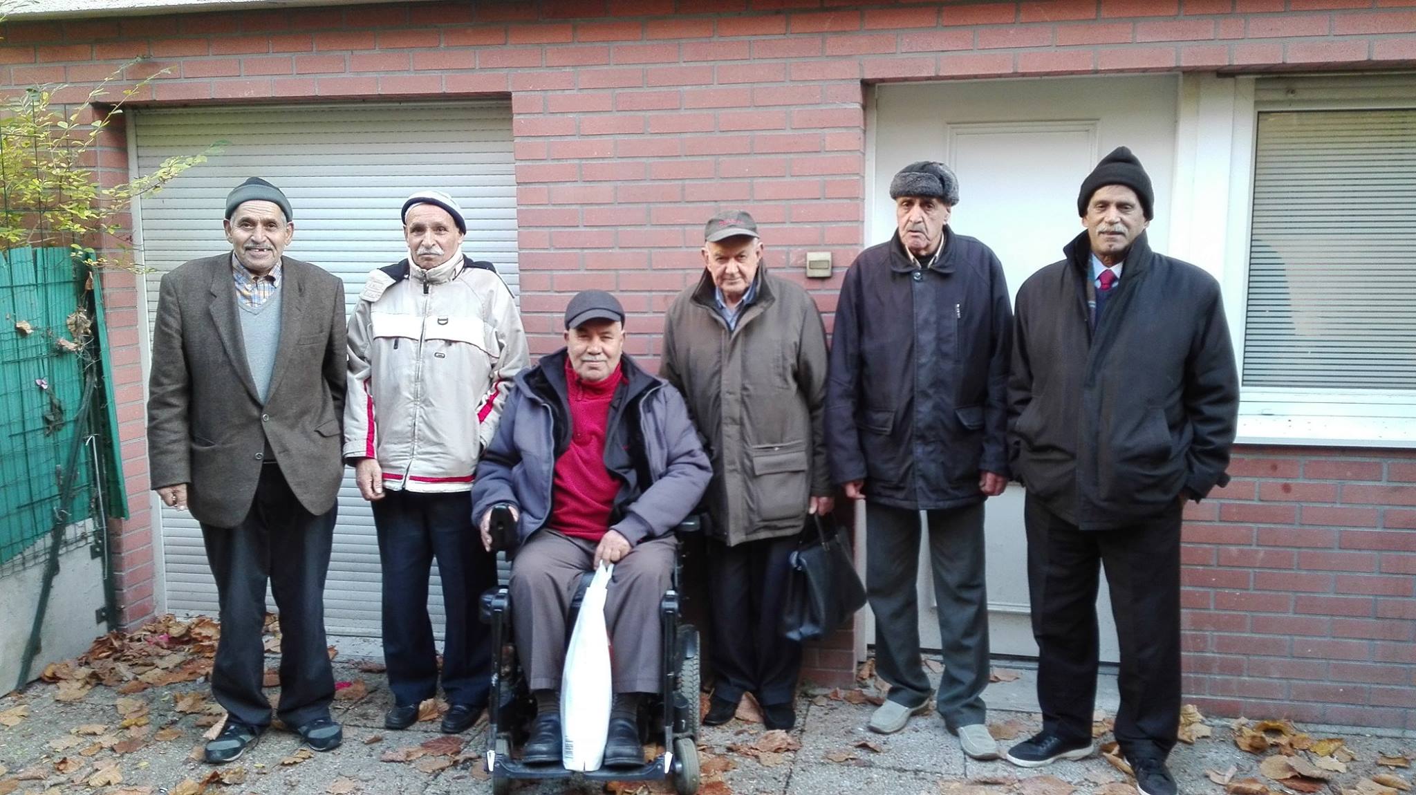 Six hommes âgés, issus de l'immigration, posent sur la terrasse de l'appartement où trois d'entre eux vient en colocation. L'un deux est en fauteuil roulant. Ce sont des amis qui fréquentent le café social Ayyem Zamen