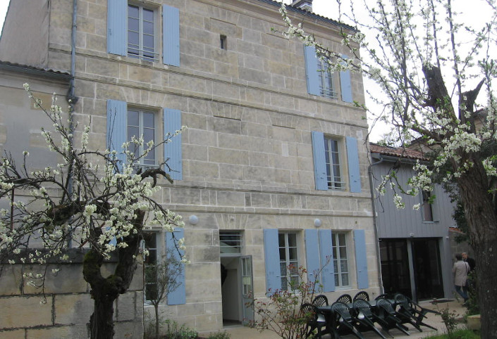 La Maison de L'Arche à Cognac, vue côté jardin. Une façade de pierres blanches de deux étages, aux volets bleus. Une grande terrasse encadrée d'arbres en fleurs. Une baie vitrée qui ouvre sur le jardin