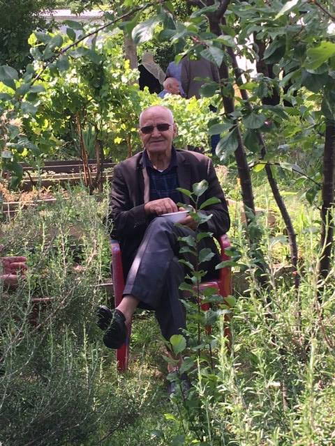 Un homme âgé issus de l'immigration est assis dans un fauteuil au milieu des herbes hautes dans un jardin . C'est un moment de détente estivale, partagé lors d'une sortie avec le café social Ayyem Zamen""