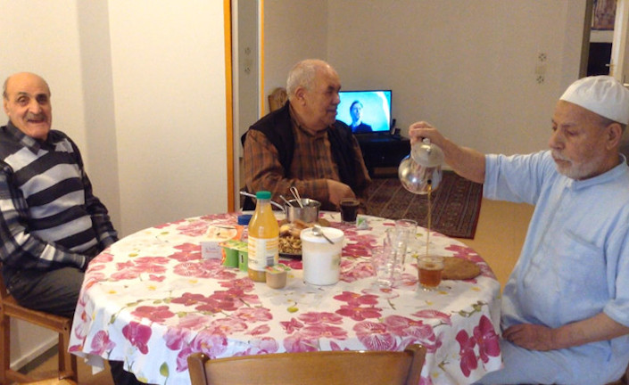 trois hommes âgés, issus de l'immigration sont assis autour d'une table dans l'appartement qu'ils habitent en colocation. L'un deux sert du thé à la menthe. Un autre à un verre de café devant lui. en arrière plan, la télévision est allumée