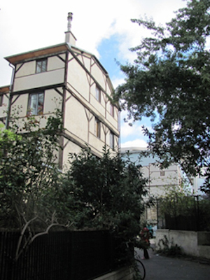 Vu du bâtiment en contre plongé. Au premier plan des arbres et une petite rue pavée. Au second plan un batiment à colombages de trois étages. 