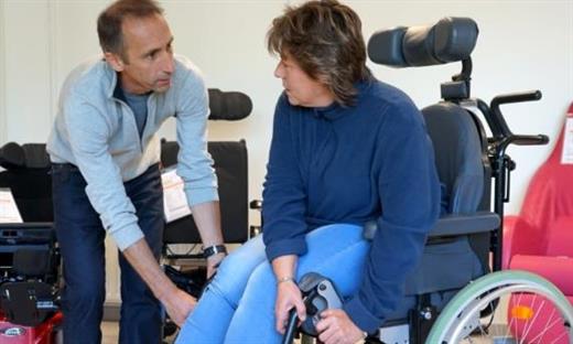 Un homme est penché vers une femme en fauteuil roulant. Il saisit quelque chose sur le côté du fauteuil en lui parlant. Les deux se regardent. 