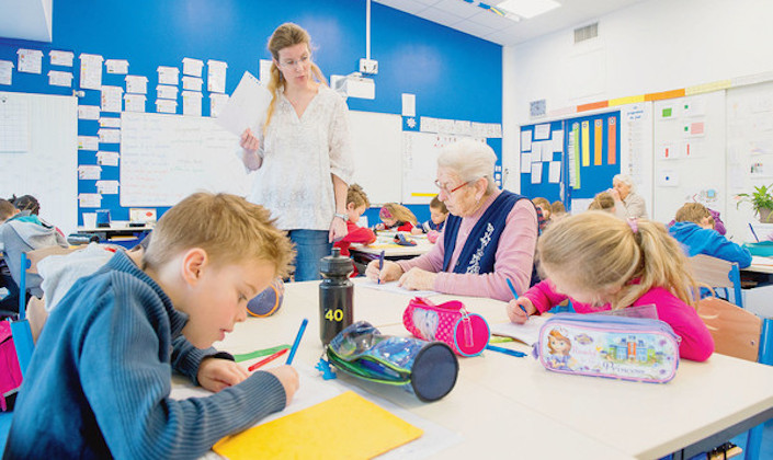 Une salle de classe aux murs bleus. Au premier plan, un table carré de quatre places. De chaque côté deux personnes sont assises : deux enfants d'une huitaine d'années d'un côté, une petite fille et une dale âgée de l'autre. Tous écrivent. Des troussent d'écoliers reposent sur la table. Au second plan, debout devant la table, l'institutrice tient une feuille à la mai et s'adresse à la dame âgée 