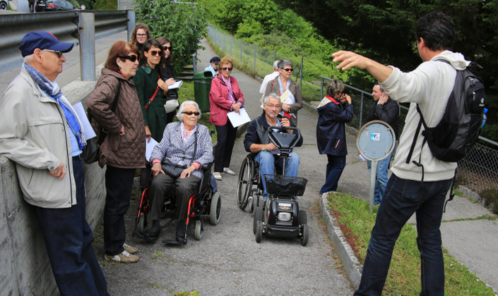 A l'exterieur, au bord d'une route, sur un chemin pieton dans ce qui semble être un parc un homme de dos parle à un groupe aligné devant lui. Dans ce groupe : deux personnes en fauteuil roulant, des seniors et des plus jeunes. 