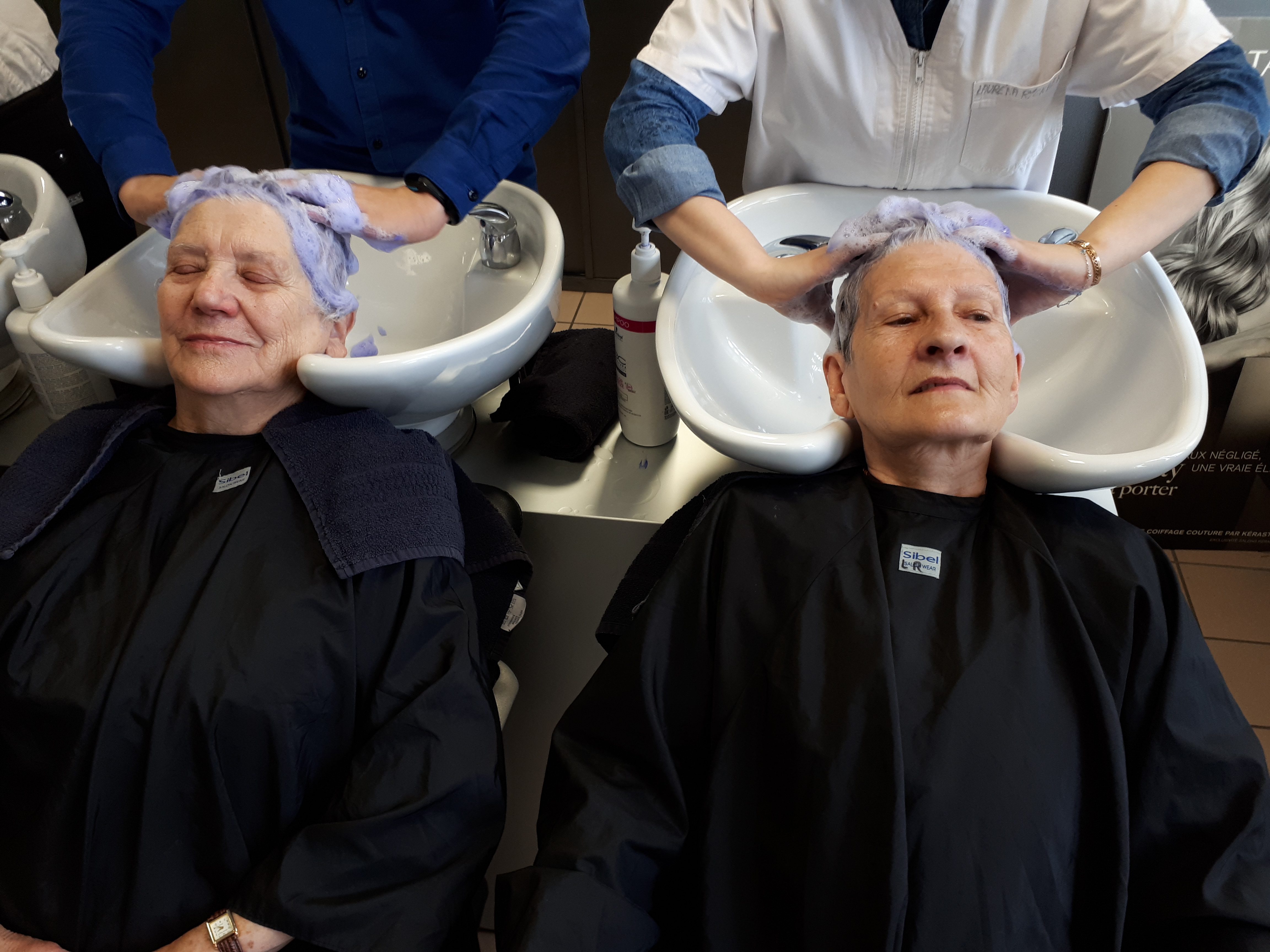 Deux femmes âgées chez le coiffeur, assises en blouse noires, les yeux clos, se font shampouiner. En arrière plan on aperçoit les mains des coiffeuses massant leur cuir cheveulu.   