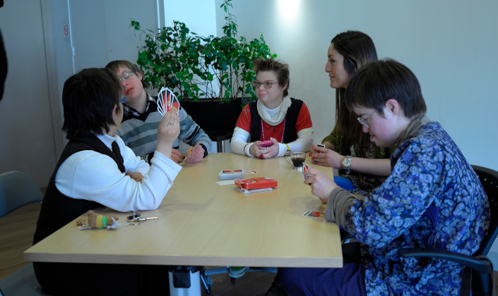 Autour d'une table, quatre jeunes adultes trisomiques et une animatricent jouent aux cartes.